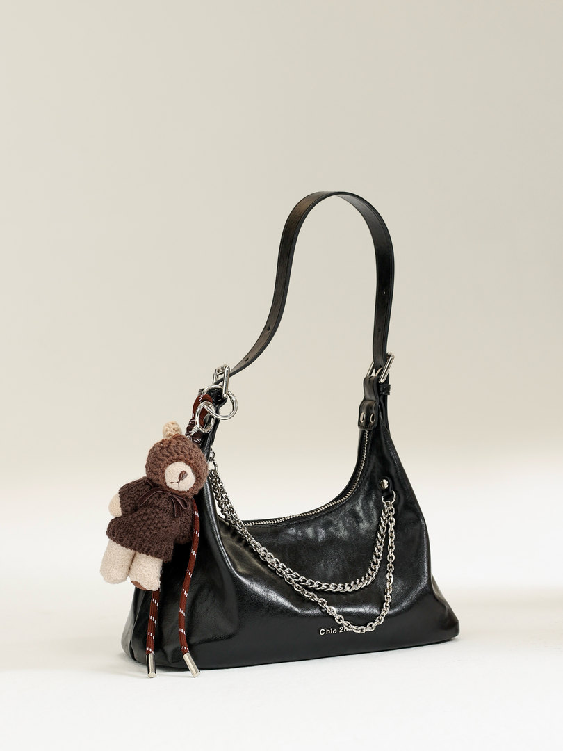 Black leather shoulder handbag with chain details and teddy bear charm on a plain background