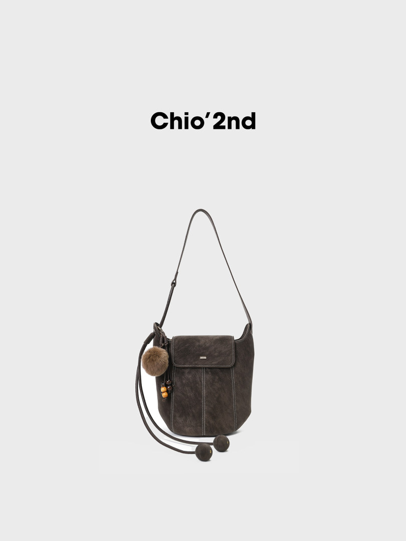 Dark brown shoulder bag with long strap and pom-pom charm on a white background