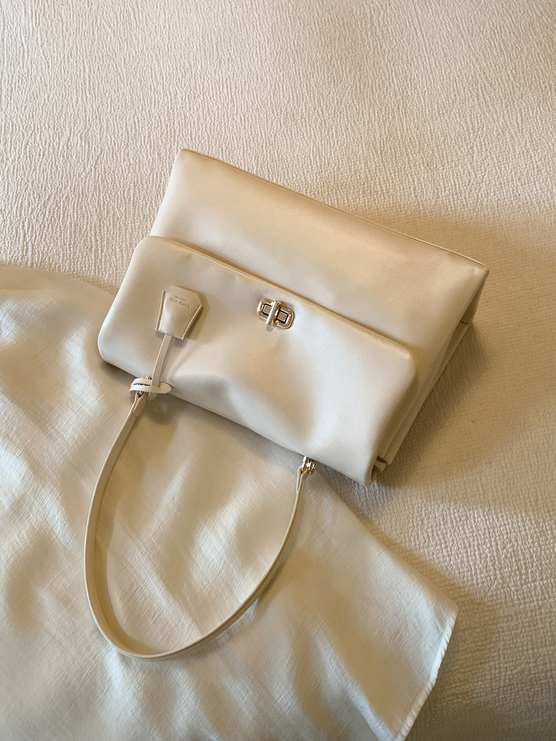 Cream-colored leather handbag with gold clasp and shoulder strap on a textured fabric surface
