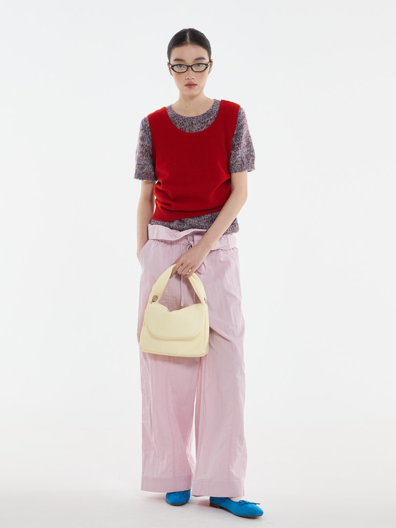 Fashion model wearing a red sweater vest, pink wide-leg pants, and holding a cream handbag
