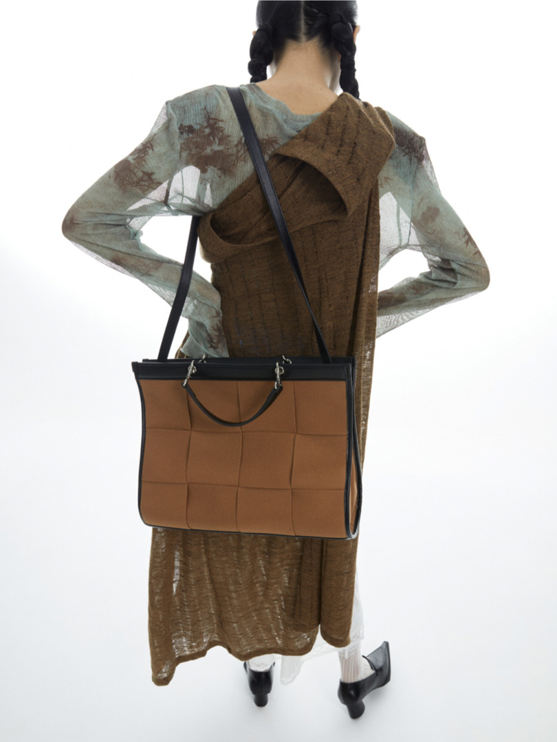 Model wearing a sheer layered brown dress with a tan tote bag