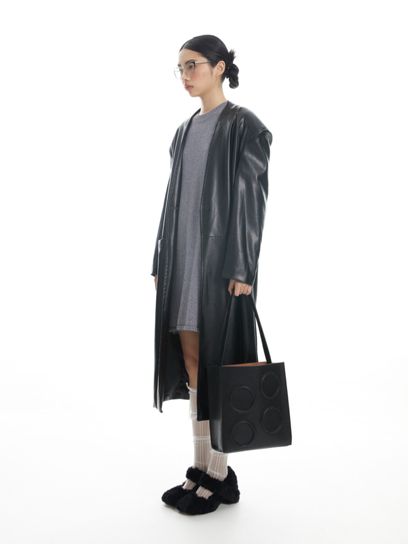 Model wearing a long black leather coat over a gray dress with a black tote bag