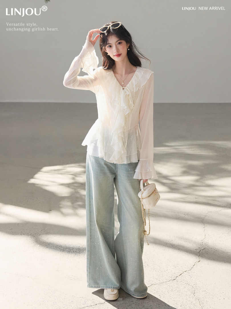 Model wearing a sheer white ruffled blouse with wide-leg light blue pants and a small handbag
