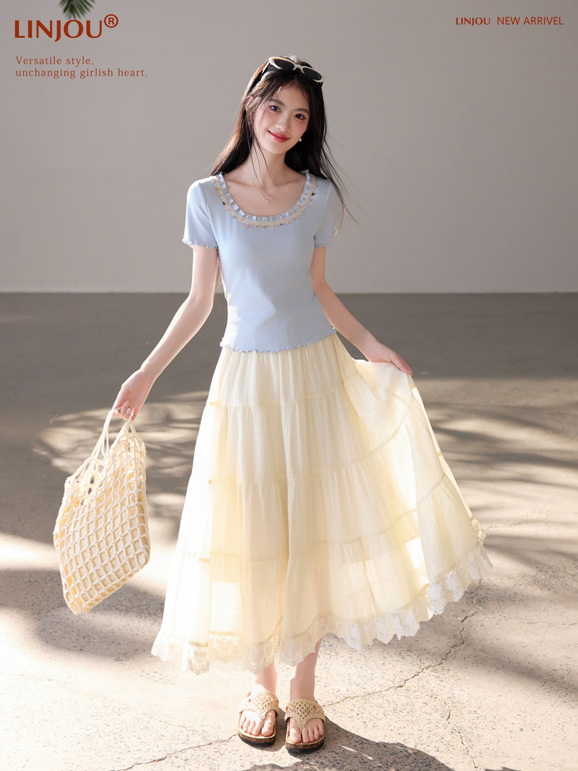 Model wearing a light blue knit top and cream tiered skirt with woven sandals and bag
