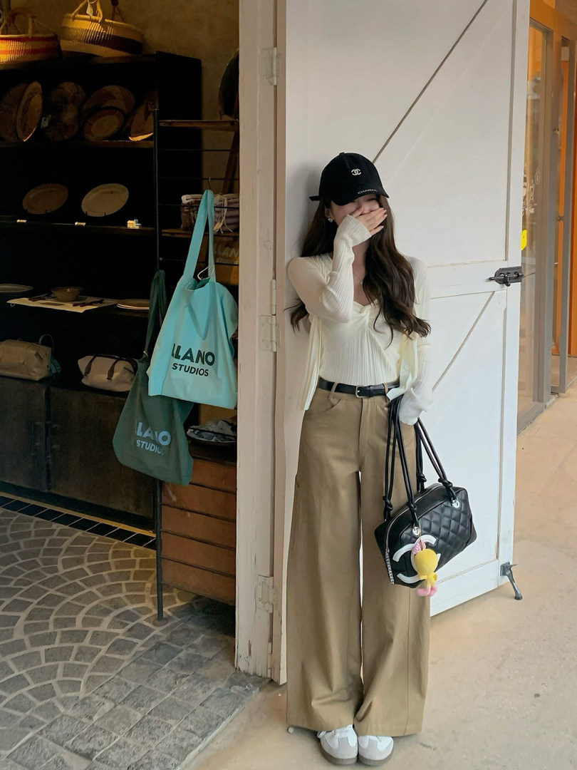 Woman wearing a cream knit top and beige wide-leg pants with a black cap and handbag