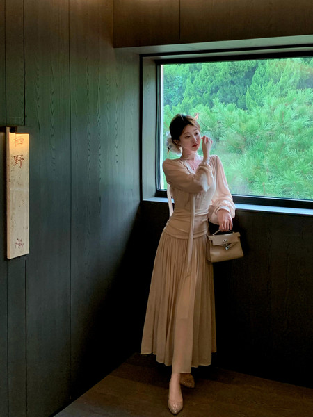 Woman wearing a beige long-sleeve pleated dress while holding a small handbag