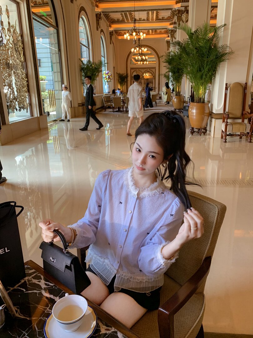 Woman wearing a light lavender frilled blouse while holding a black handbag in a hotel lobby