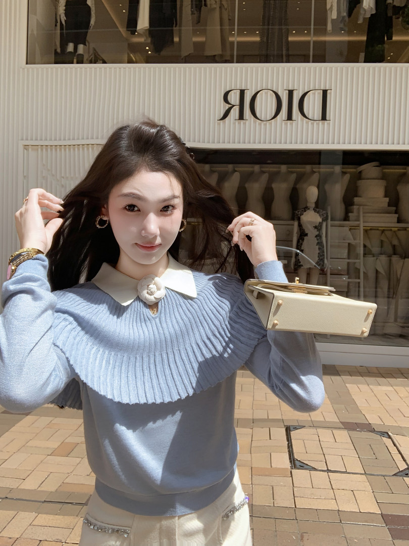 Woman wearing a light blue ribbed sweater with a white collar and flower detail, holding a cream handbag.