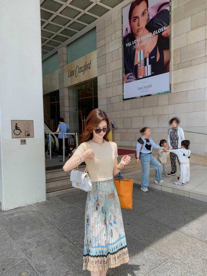 Woman wearing a cream top and pastel pleated skirt with a white quilted handbag