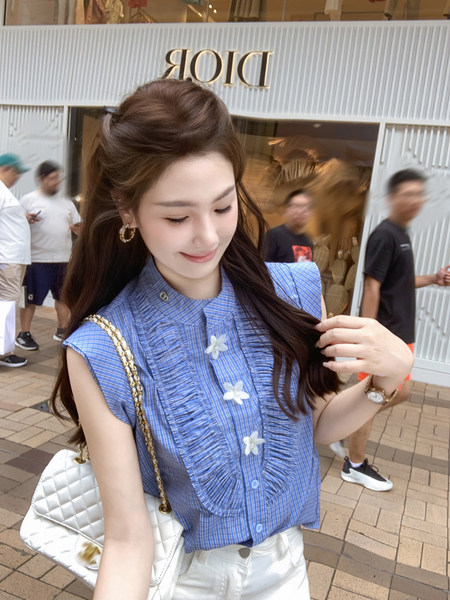Woman wearing a blue sleeveless ruffled blouse with white floral details and a white quilted handbag