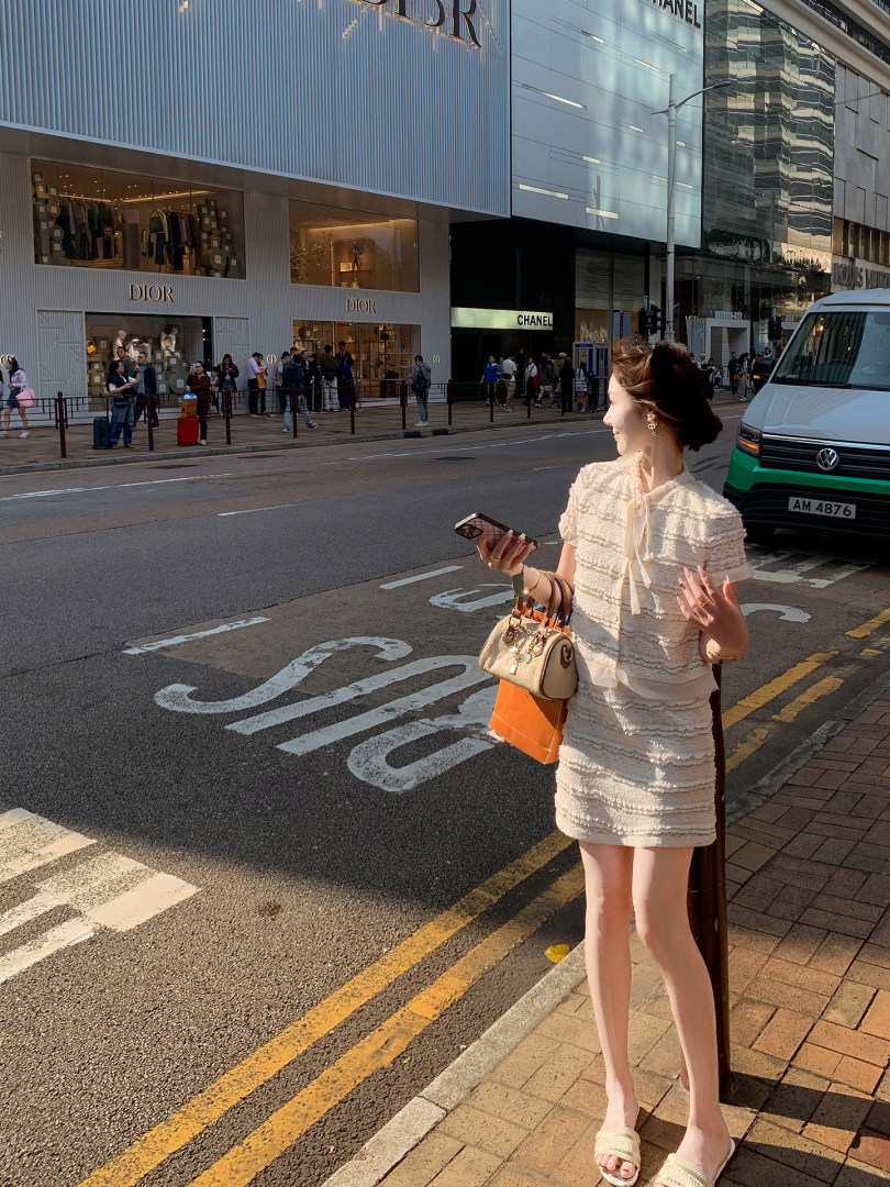Woman wearing a cream textured short dress and sandals while holding a handbag on a city street