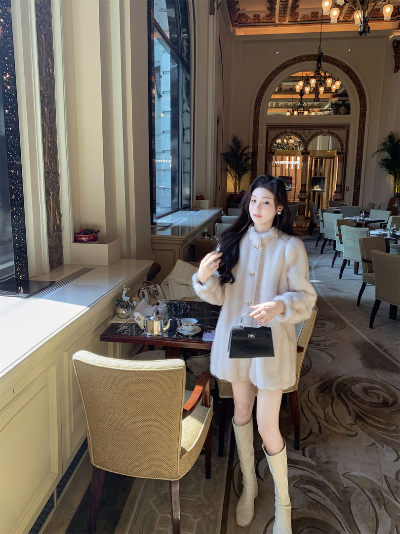 Woman modeling a cream coat dress with boots and a black handbag in a luxury restaurant