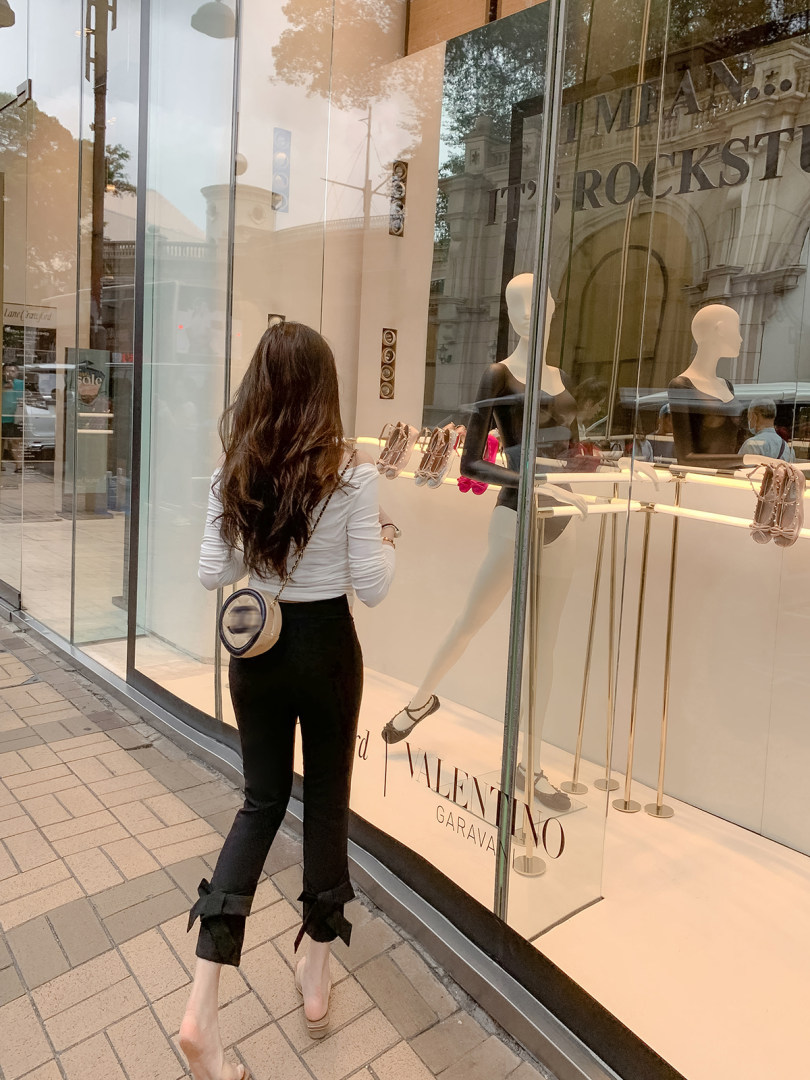 Woman wearing a white top and black bow-hem pants with a small round crossbody bag outside a luxury boutique