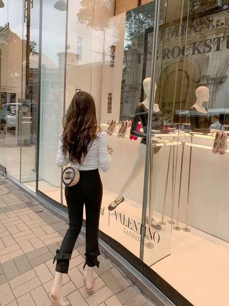 Woman wearing a white top and black bow-hem pants with a small round crossbody bag outside a luxury boutique