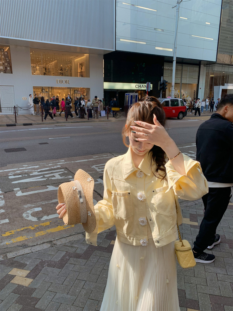 Woman wearing a pale yellow jacket and cream pleated dress while holding a straw hat and small handbag on a city street