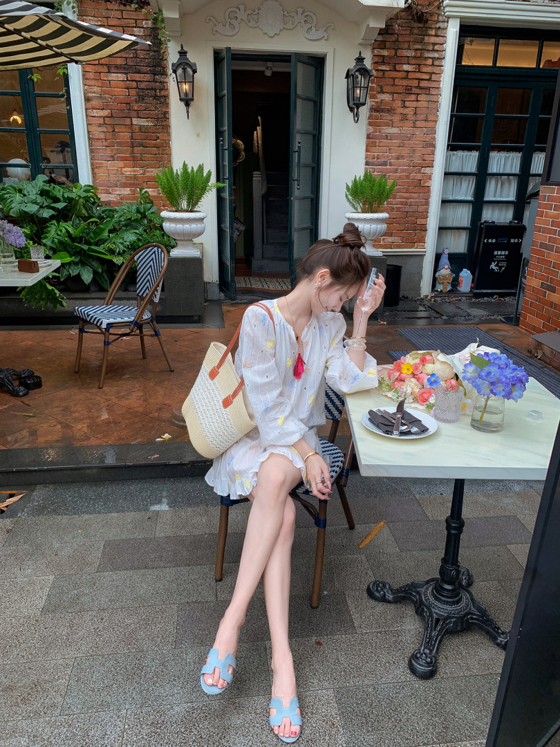 Woman wearing a white floral dress with a straw tote bag and blue sandals at an outdoor cafe