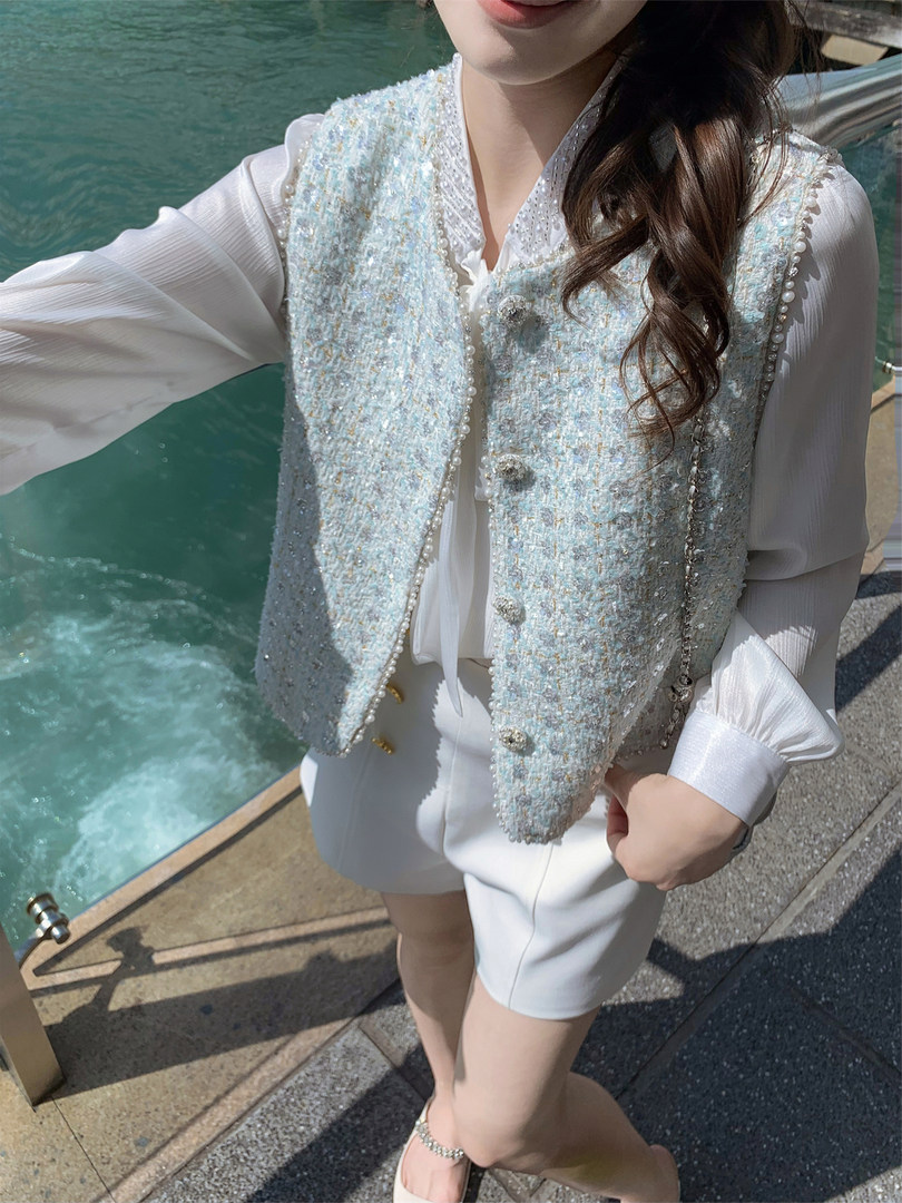 Woman wearing a light blue tweed vest over a white outfit by the water