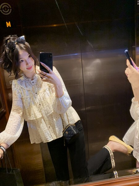 Mirror selfie of a woman wearing a white ruffled blouse and black pants with a small black quilted handbag