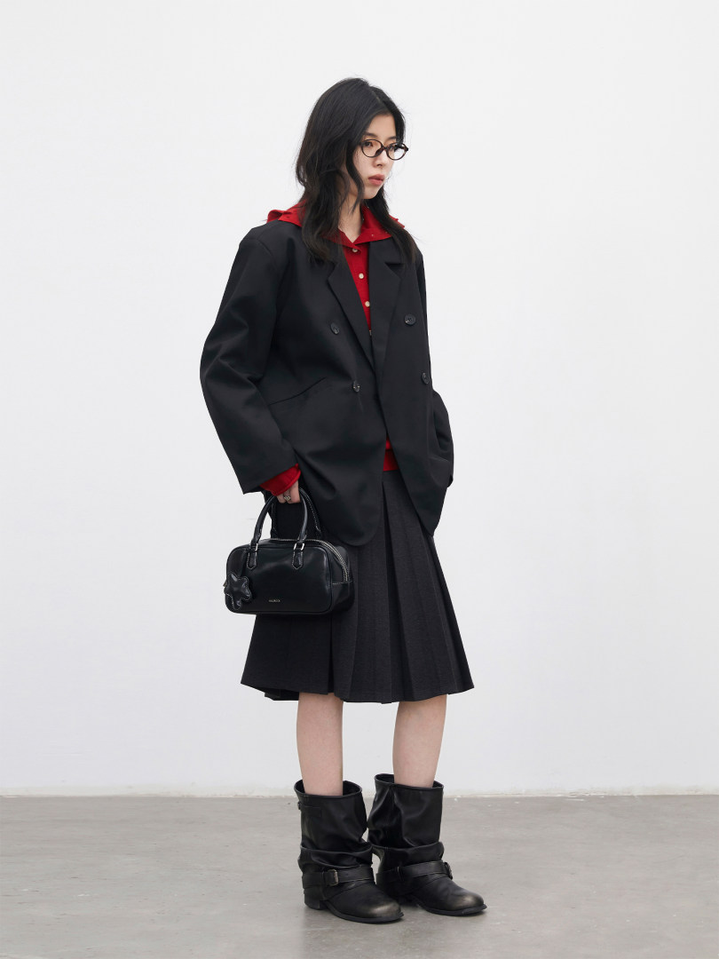 Model wearing a black blazer, pleated skirt, boots, and carrying a black handbag