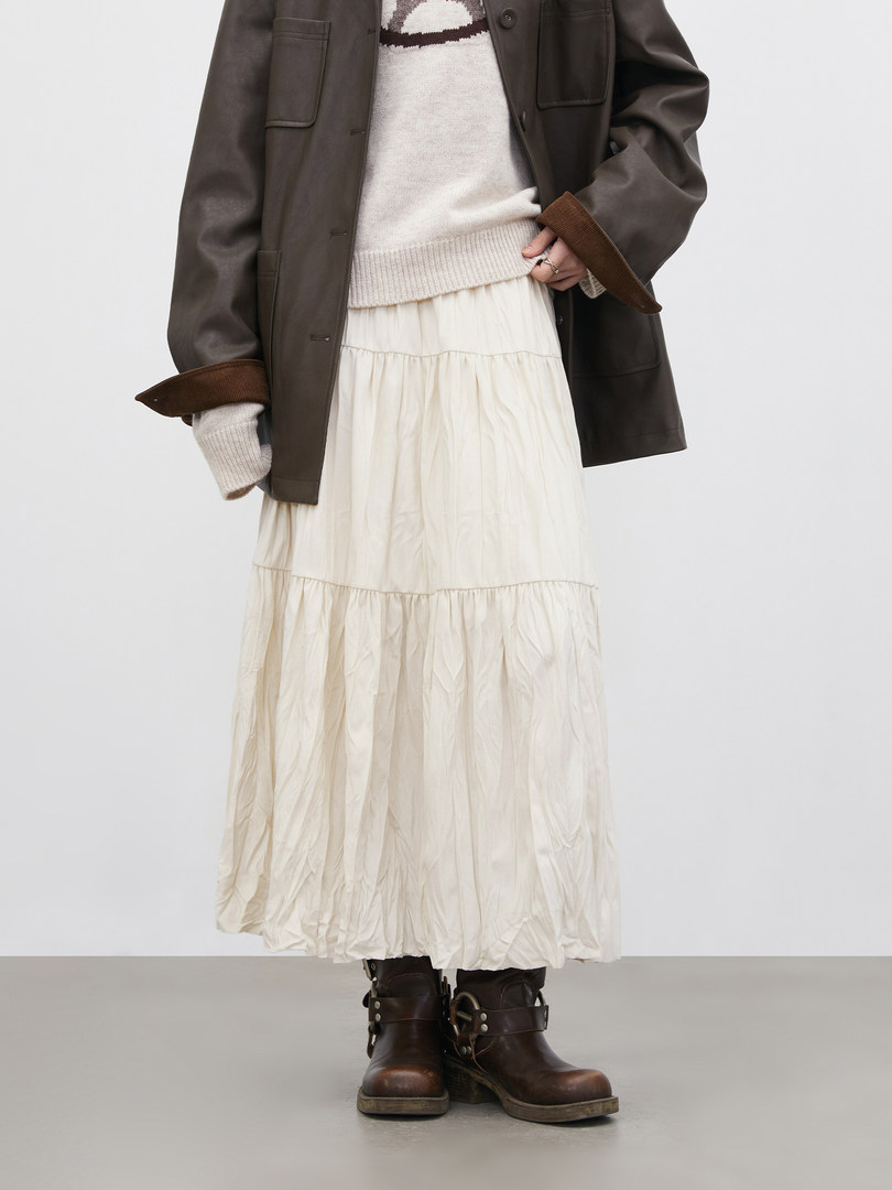 Model wearing a layered outfit with dark jacket, cream sweater, long white skirt, and brown boots
