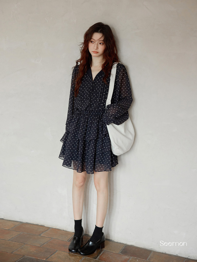Model wearing a black polka dot long-sleeve mini dress with a white shoulder bag