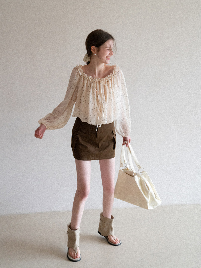 Woman wearing a cream sheer blouse, brown mini skirt, sandals, and holding a beige tote bag