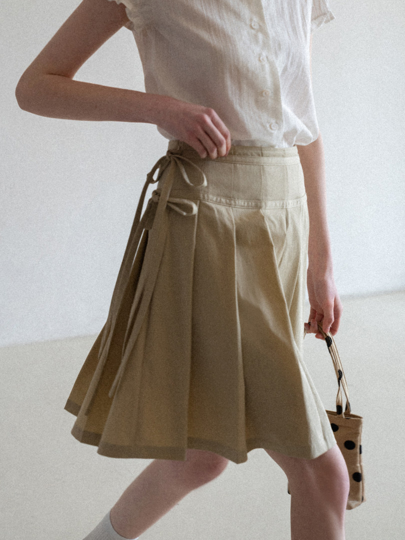 Model wearing a light beige pleated skirt with a white button-up top and a small handbag