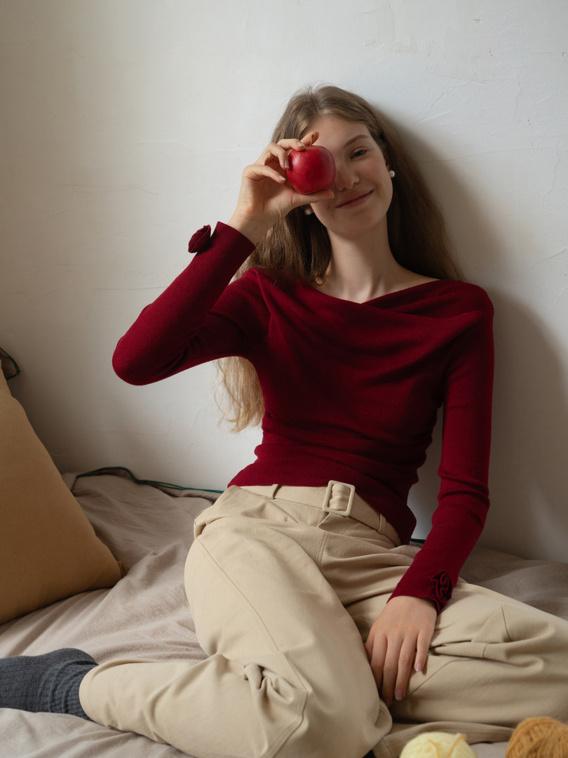 Woman wearing a burgundy long-sleeve top and beige trousers while holding a red apple