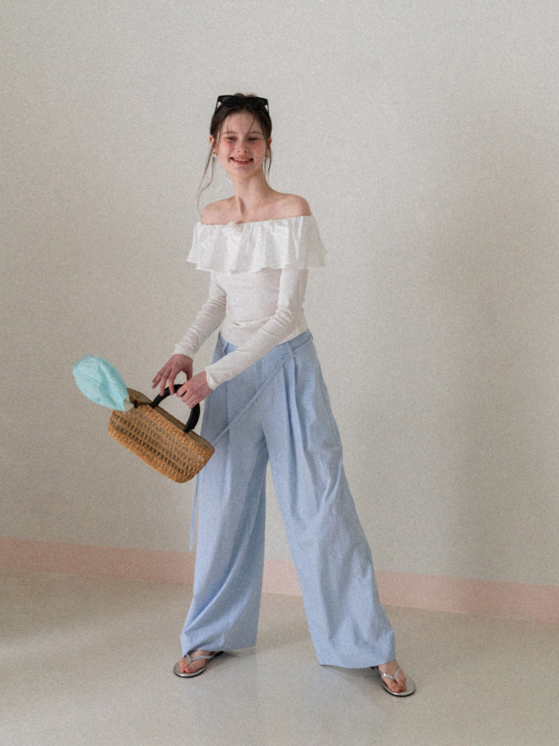 Model wearing a white off-shoulder top with light blue wide-leg pants and sandals, holding a woven bag