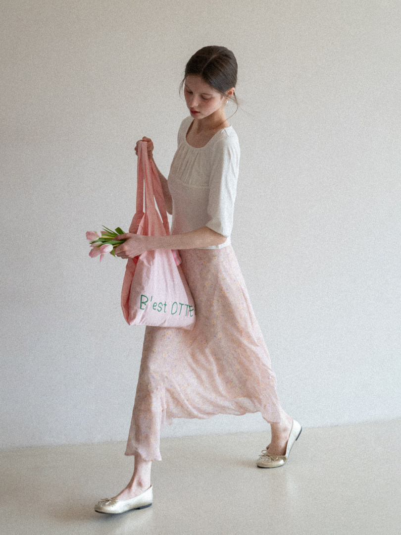 Woman wearing a white top and pink floral skirt while holding a pink tote bag and flowers