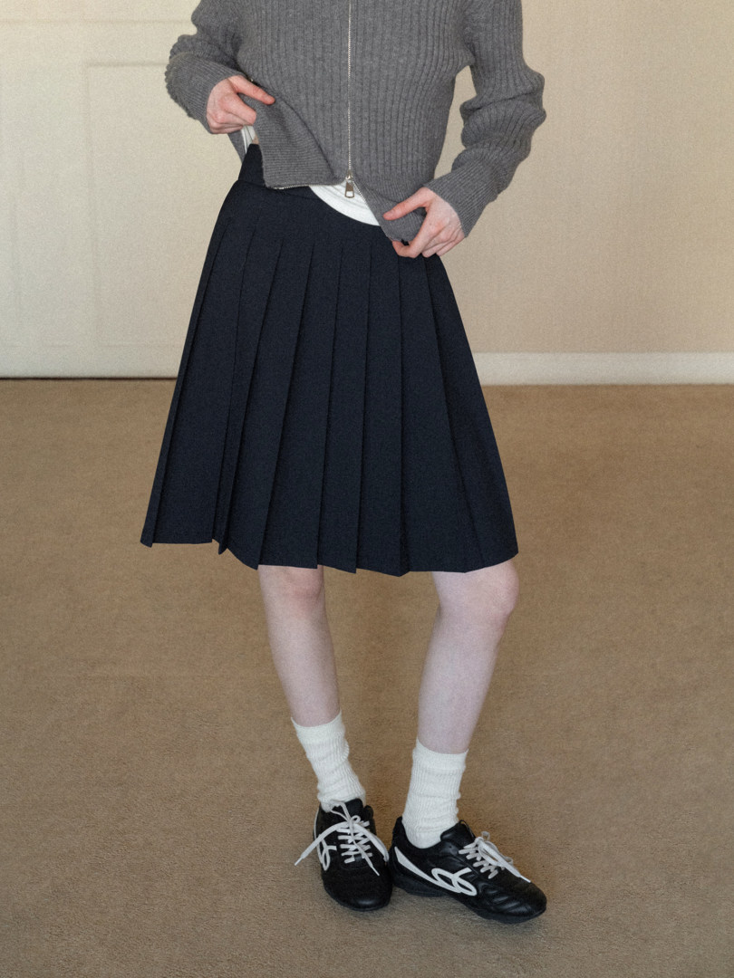 Model wearing a black pleated skirt with a gray knit zip sweater and black sneakers