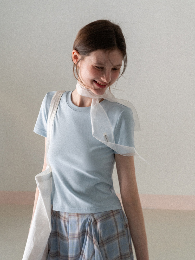 Model wearing a light blue short-sleeve top with a plaid skirt and sheer white scarf
