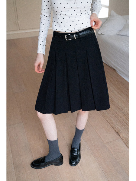 Model wearing a black pleated skirt with belt in a bedroom setting