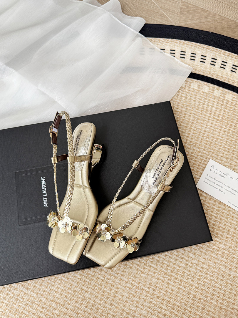 Gold heeled sandals with braided straps and floral embellishments displayed on a box