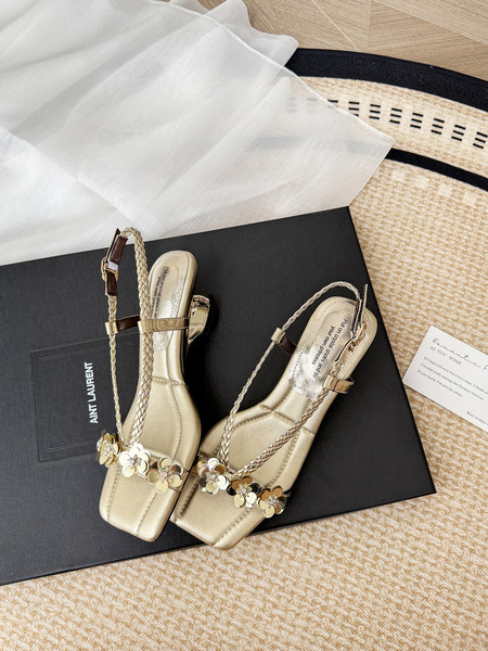 Gold heeled sandals with braided straps and floral embellishments displayed on a box