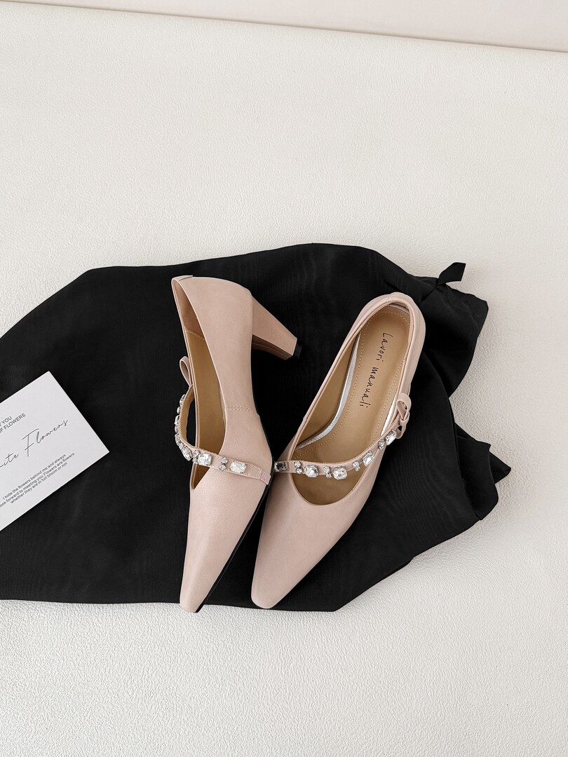Pair of nude pointed-toe high heels with rhinestone strap displayed on a black cloth