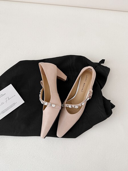 Pair of nude pointed-toe high heels with rhinestone strap displayed on a black cloth