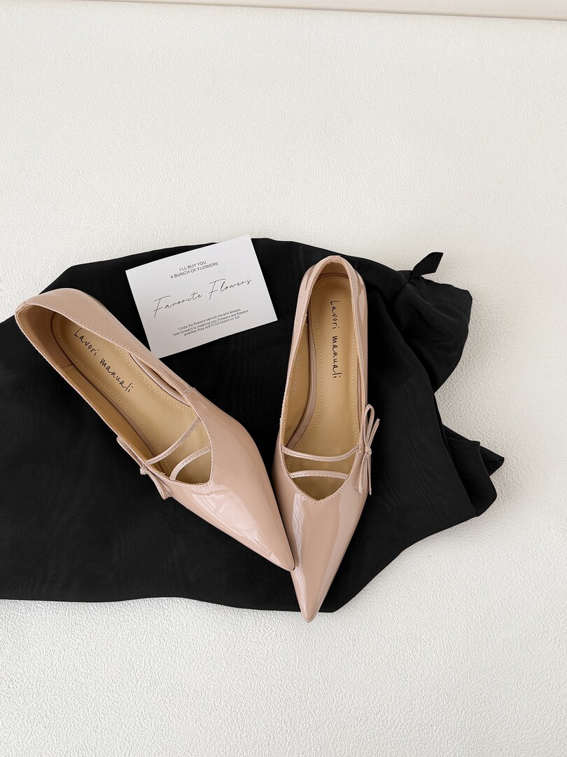 Pair of nude pink pointed-toe flats displayed on black fabric with a small branded card