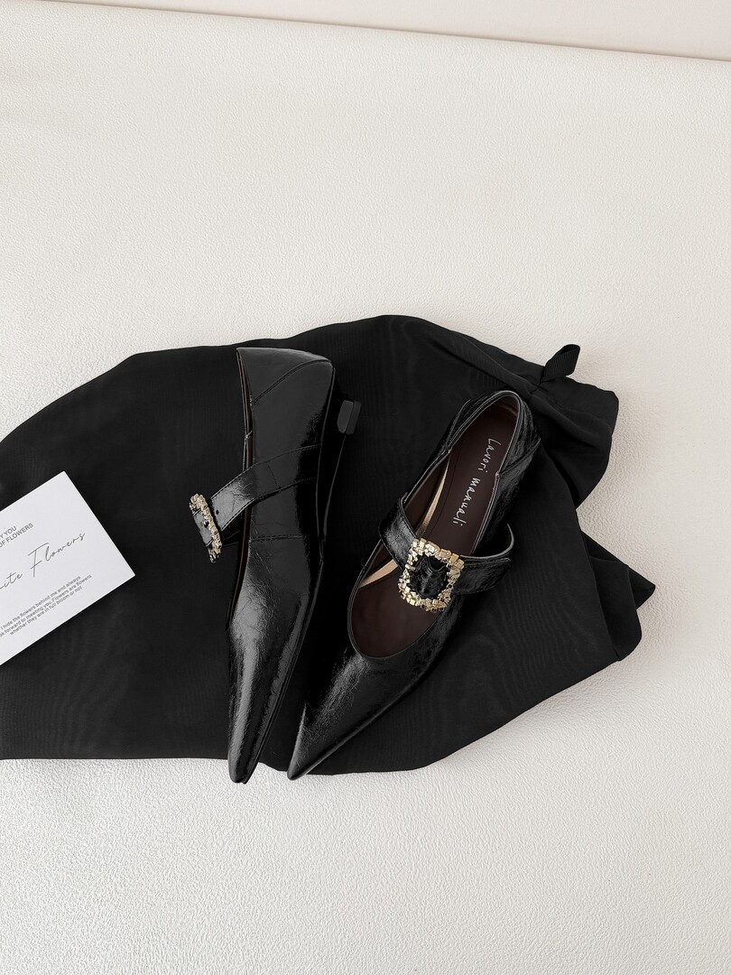 Black pointed-toe heeled flats with gold buckle detail on a black fabric background