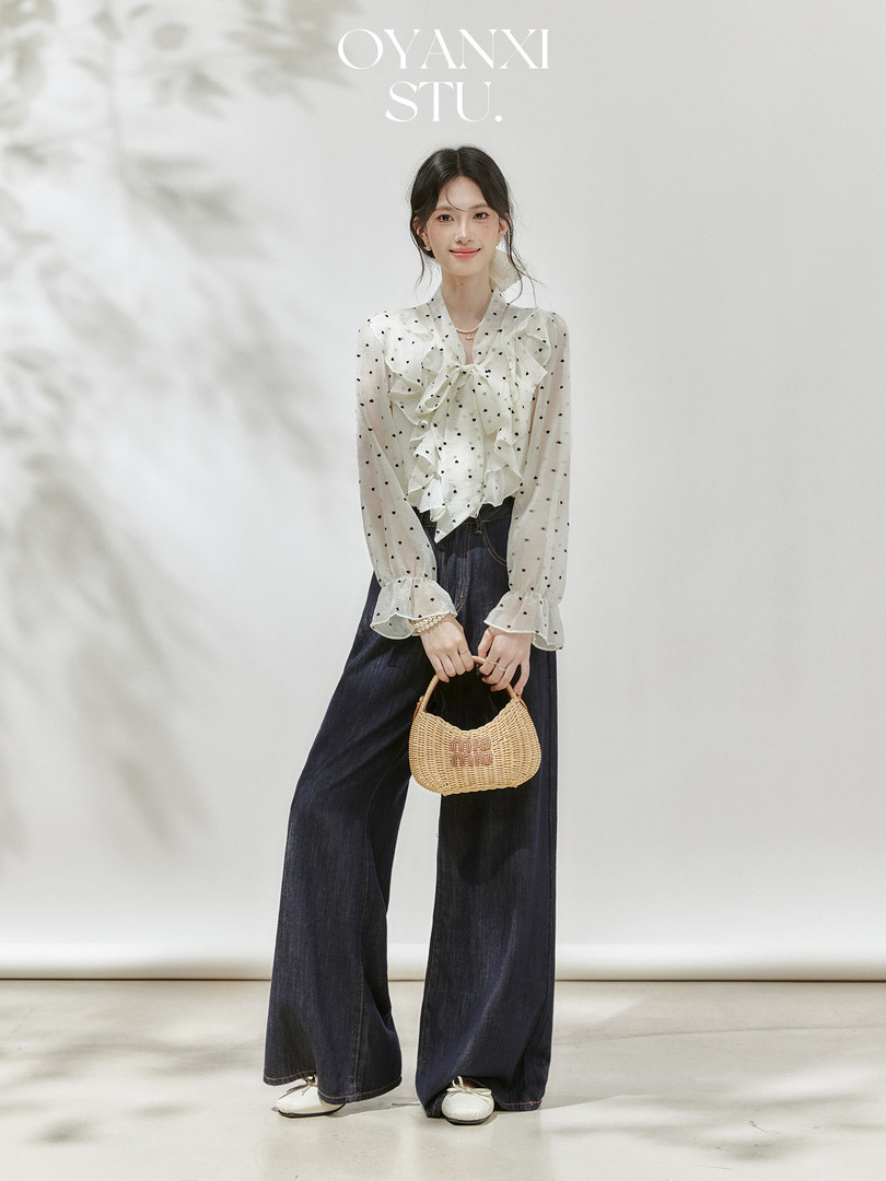 Model wearing a sheer polka dot blouse with dark wide-leg jeans and a woven handbag
