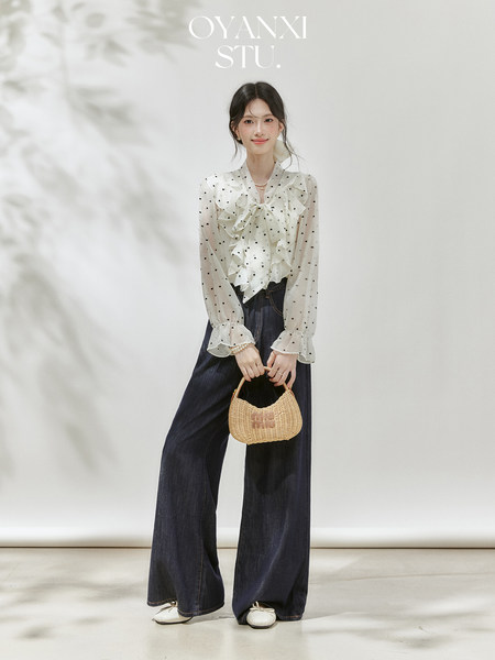 Model wearing a sheer polka dot blouse with dark wide-leg jeans and a woven handbag