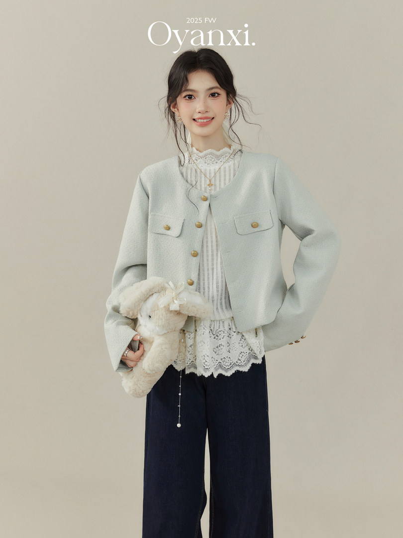 Model wearing a light gray cropped jacket over a lace blouse with wide-leg dark pants, holding a plush bunny accessory