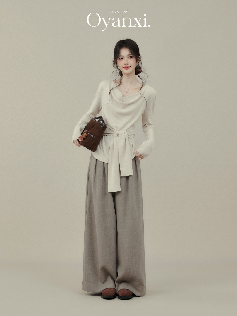 Model wearing a cream knit top and wide-leg gray pants with a brown handbag