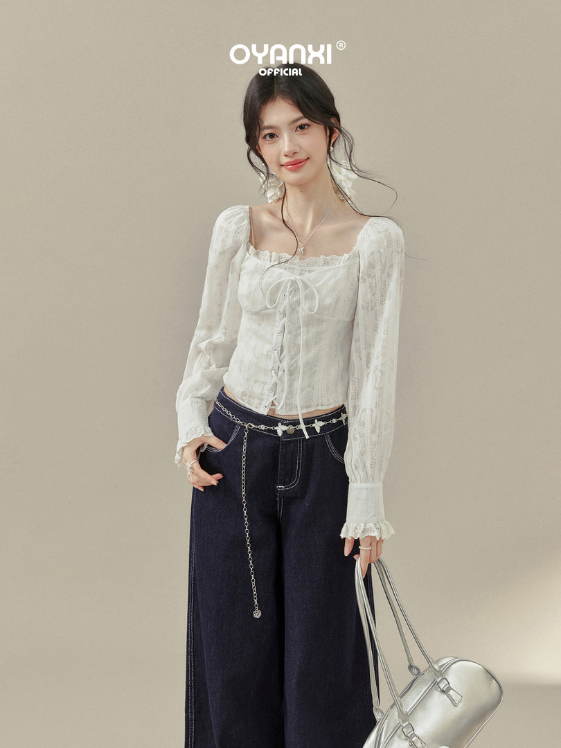 Model wearing a white lace corset-style blouse with dark wide-leg jeans and a silver handbag