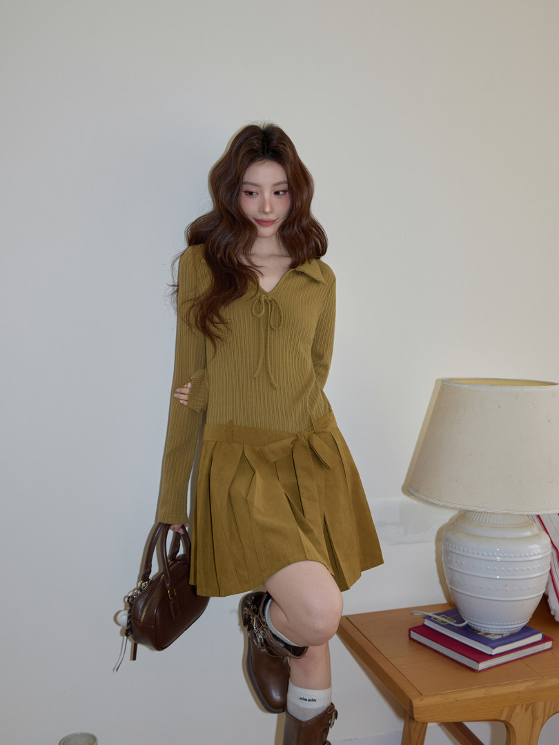 Model wearing an olive ribbed long-sleeve top and matching pleated mini skirt with boots and handbag