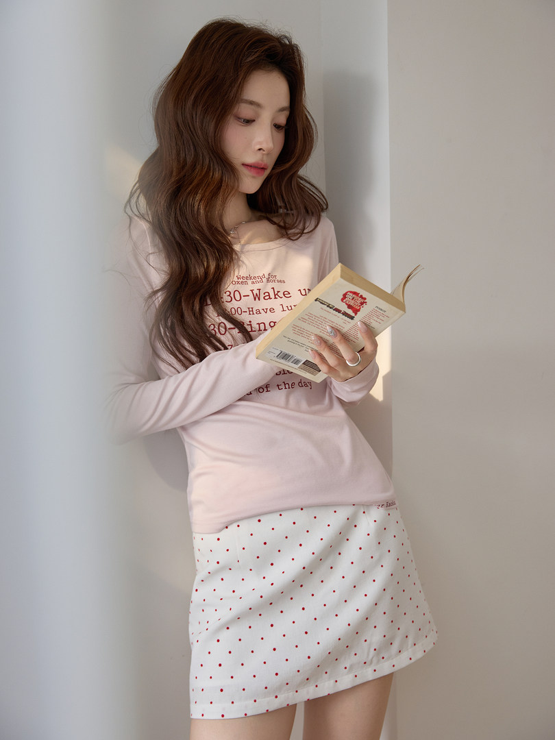 Model wearing a pink long-sleeve top and white polka dot skirt while reading a book