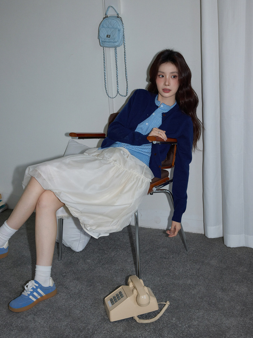 Model wearing a blue cardigan, striped top, white skirt, and blue sneakers with a small backpack