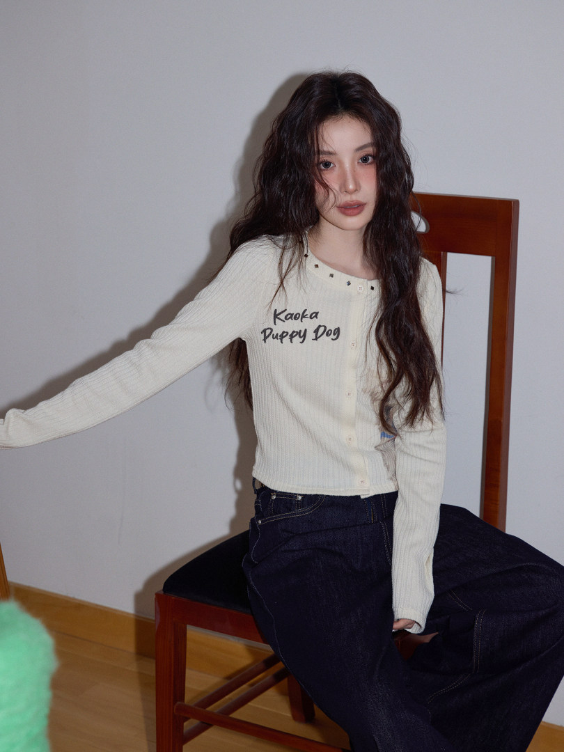Woman wearing a cream long-sleeve top and dark jeans while seated on a chair