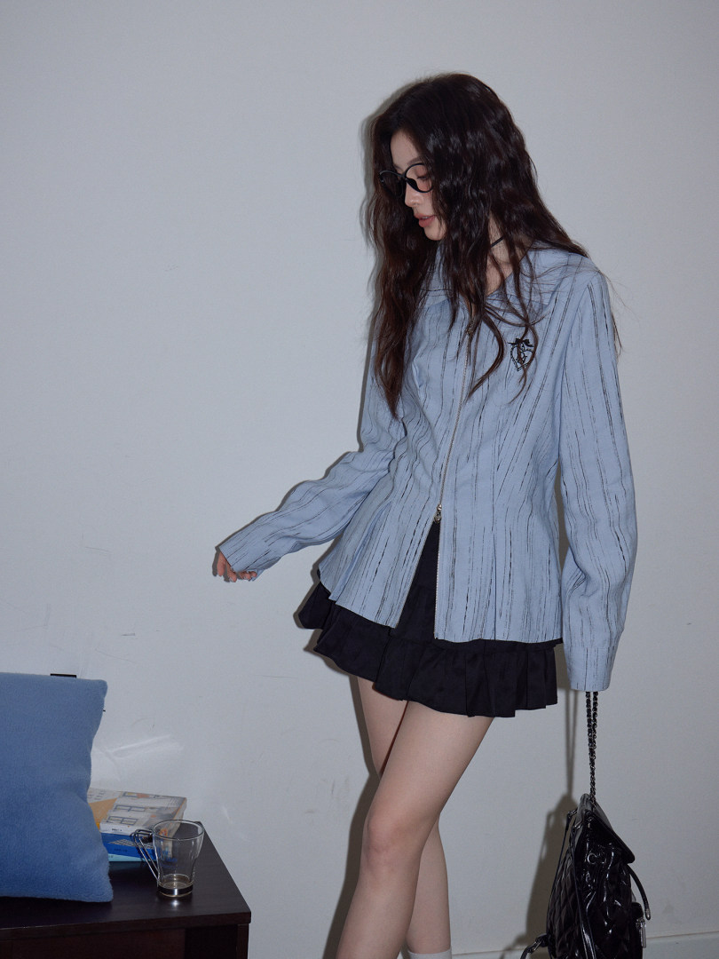 Model wearing a light blue zip-front long-sleeve top with a black pleated skirt