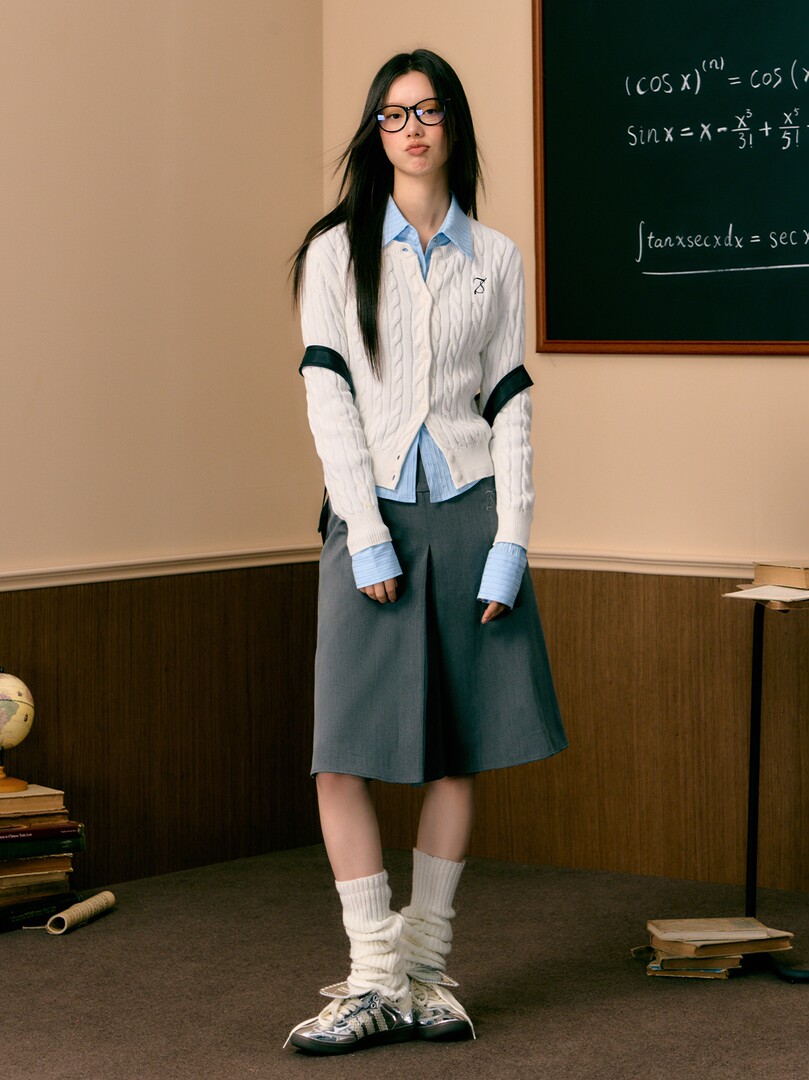 Model wearing a preppy school-style outfit with a white cable-knit cardigan, blue shirt, gray skirt, and sneakers